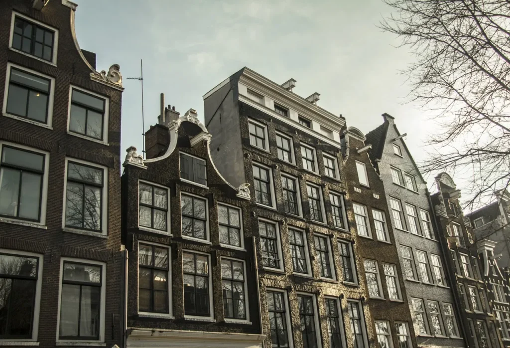 Traditional tall, narrow canal houses with characteristic gabled roofs line a street in Amsterdam, Netherlands, under a bright sky.