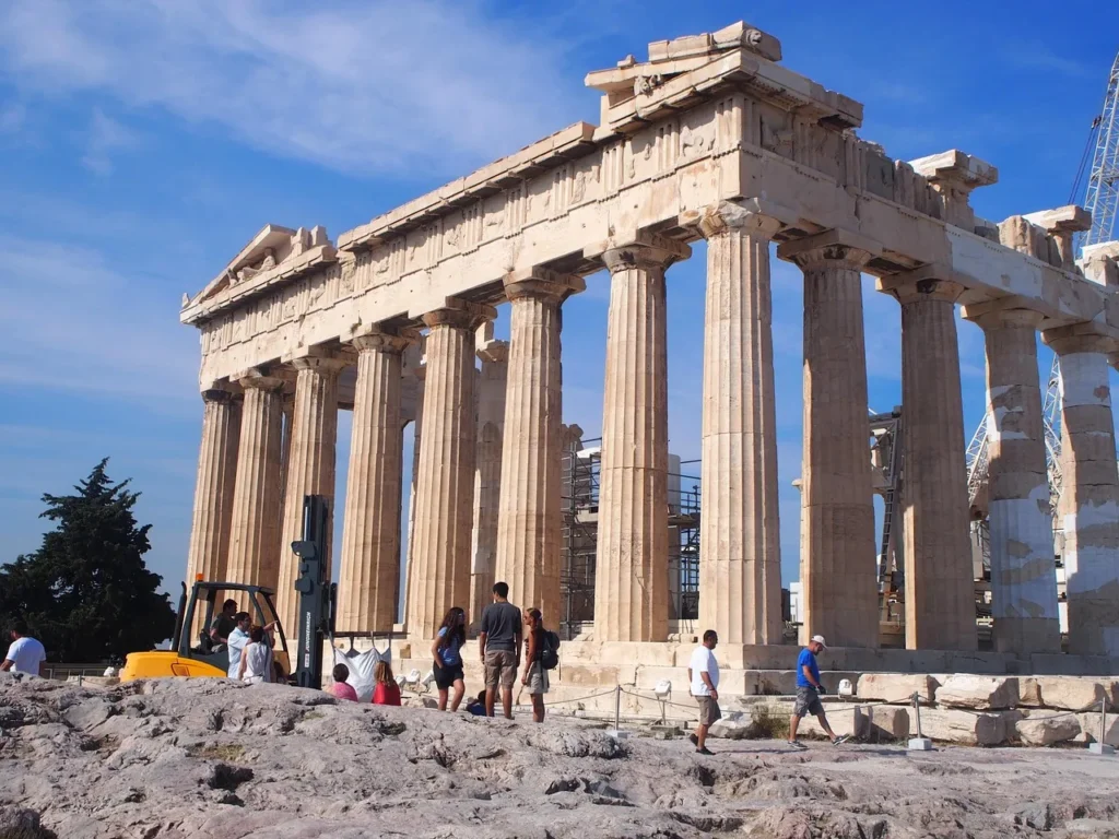 The ancient Parthenon temple, a large stone structure with tall columns, standing under a blue sky with some people walking around its rocky base in Athens, Greece.