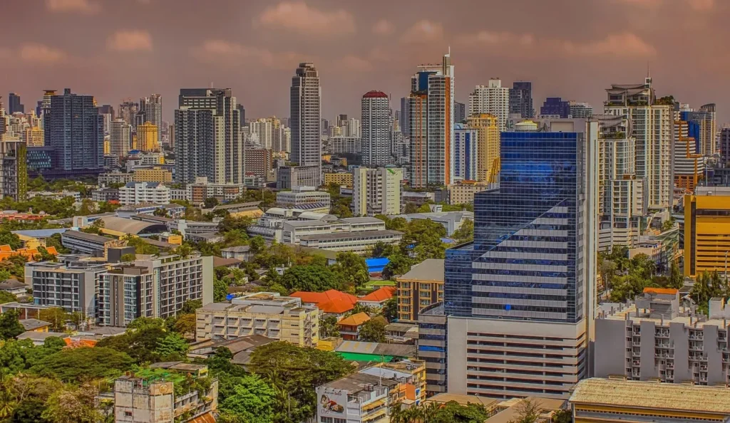 An aerial view of the Bangkok skyline, featuring a dense cluster of modern high-rise buildings and skyscrapers interspersed with green trees, under an overcast sky.