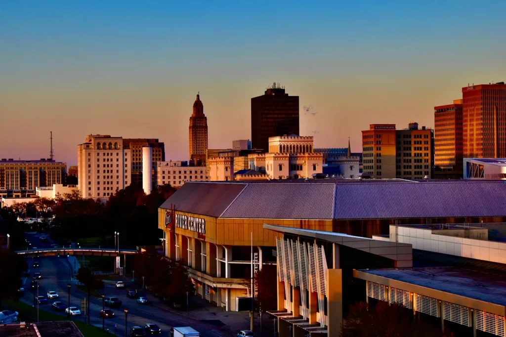 An elevated view of the Baton Rouge skyline at dusk, showing various buildings including the State Capitol building with its distinctive tower, and the Baton Rouge River Center in the foreground, under a sky with hues of orange and blue.