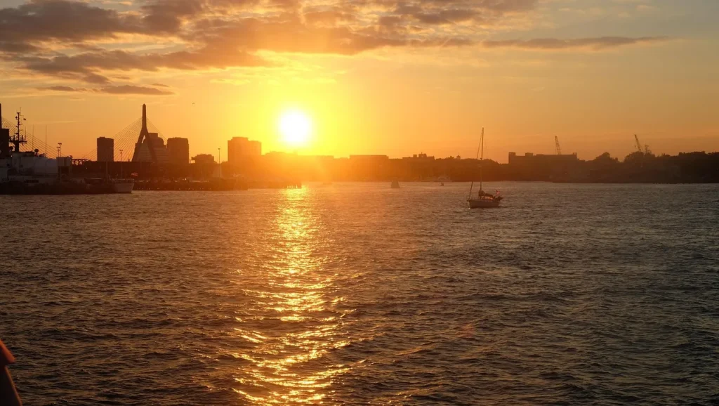 The sun setting over a body of water in Boston, casting an orange glow across the sky and reflecting on the water, with the city skyline and a bridge silhouetted in the distance.