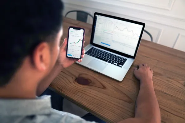 An individual reviewing financial charts on both a smartphone and a laptop, symbolizing the accessibility of market data and trading platforms offered through MyFastBroker.com.