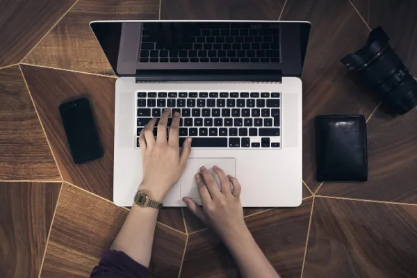 Overhead shot of hands typing on a silver laptop keyboard, with a smartphone, a camera lens, and a wallet on a wooden desk.