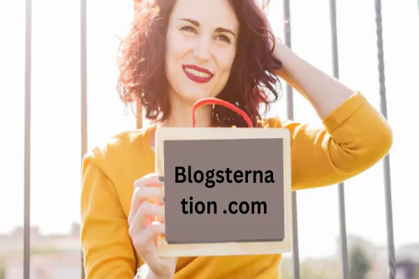A smiling woman with red curly hair and red lipstick, wearing a mustard yellow top, holding up a small chalkboard with "Blogsternation .com" written on it. The background is bright and out of focus, suggesting an outdoor setting.