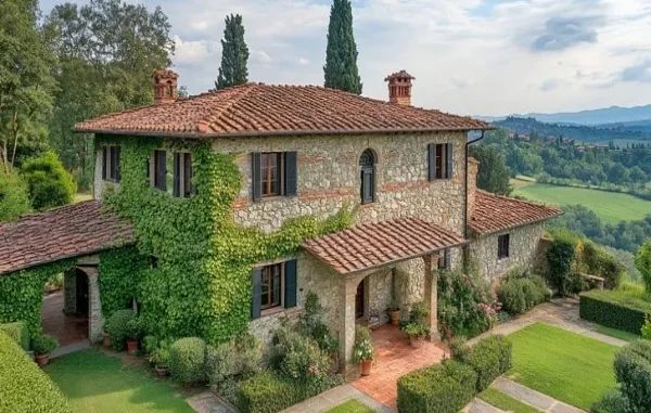 A picturesque Italian country house with stone walls and a terracotta roof, partially covered in vibrant green ivy. The villa is surrounded by meticulously manicured gardens, tall cypress trees, and rolling green hills under a bright sky with scattered clouds.