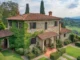 A picturesque Italian country house with stone walls and a terracotta roof, partially covered in vibrant green ivy. The villa is surrounded by meticulously manicured gardens, tall cypress trees, and rolling green hills under a bright sky with scattered clouds.