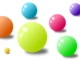 Various colorful, shiny spheres of different sizes, including green, pink, orange, blue, and purple, scattered on a white background, resembling bubblegum bubbles.