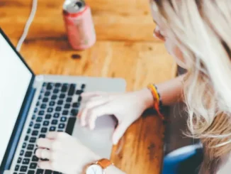 An overhead, shallow depth of field shot of a person with blonde hair typing on a silver laptop keyboard on a wooden table, with a pink aluminum can blurred in the background, symbolizing engagement with online content.