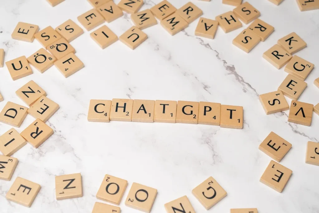 Wooden scrabble tiles spelling out the letters C.H.A.T.G.P.T. in a line on a white marble surface, surrounded by other scattered letter tiles, symbolizing the underlying language components of the AI.