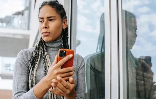 A woman with braided hair leaning against a glass window, looking away with a pained or thoughtful expression while holding an orange smartphone. Her reflection is visible in the glass.