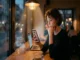 A woman sitting at a warm, lit cafe counter near a window, thoughtfully looking at a dating profile on her smartphone as subtle holographic AI conversation prompts and bio suggestions float nearby.